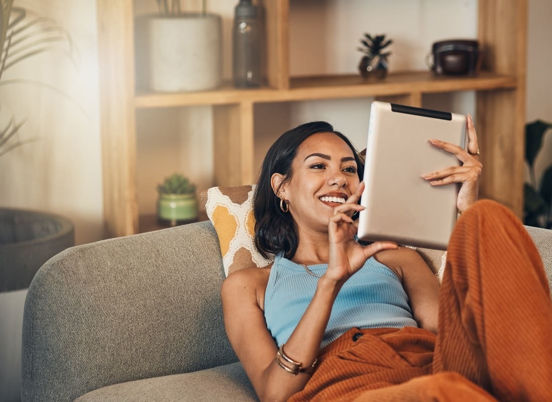Blog - Smiling Woman Relaxing On A Gray Sofa Using A Tablet In A Bright Living Room With Wooden Shelves